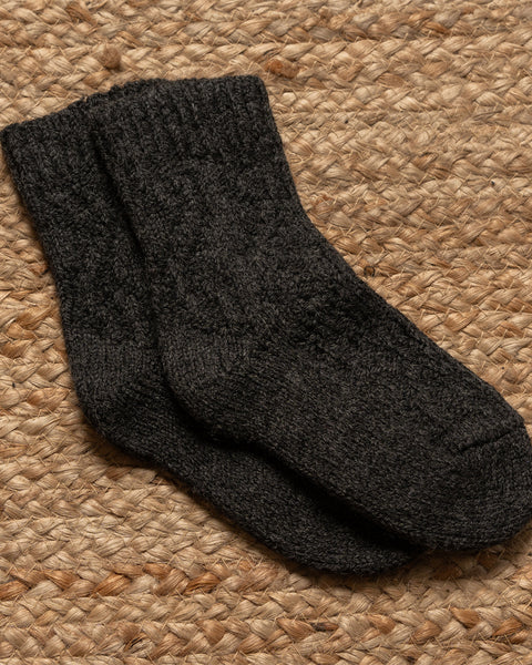 A pair of menique brand dark gray, possibly charcoal or heather black, knitted socks with a visible ribbed cuff are lying flat on a textured, light brown woven mat.