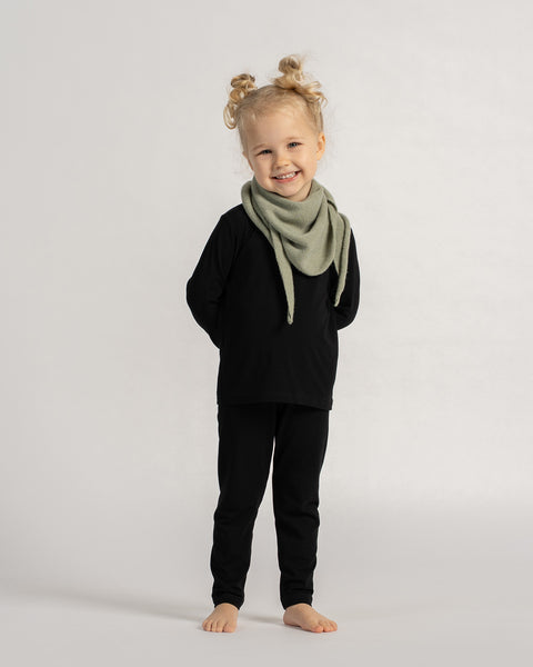 Happy child wearing a light green knit triangle scarf with a black outfit, standing barefoot against a neutral background.