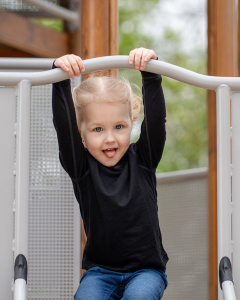 A young child with blonde hair and blue eyes, wearing a black long-sleeved shirt and blue jeans, is hanging onto a metal bar on a playground structure. They are looking directly at the camera with a happy, open-mouthed expression and their tongue slightly sticking out.
