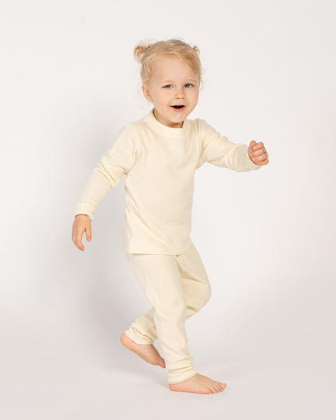 A smiling baby wearing natural merino thermal pants and a matching long-sleeve top, standing against a white background.