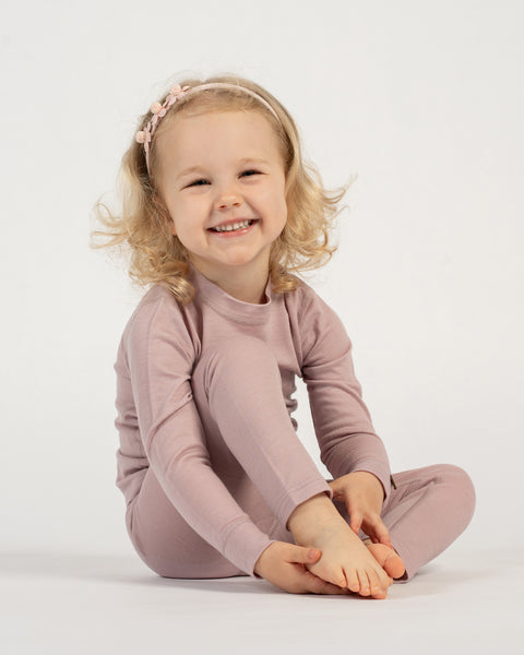 A smiling child with curly blonde hair and a light pink floral headband sits cross-legged on a white background, wearing a dusty pink long-sleeved top and matching pants. The child is looking directly at the camera.