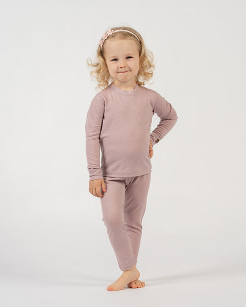 A child with blonde curly hair and a light pink floral headband stands against a white background, wearing a dusty pink long-sleeved top and matching pants. The child has one hand on their hip and is looking at the camera with a slight smile.
