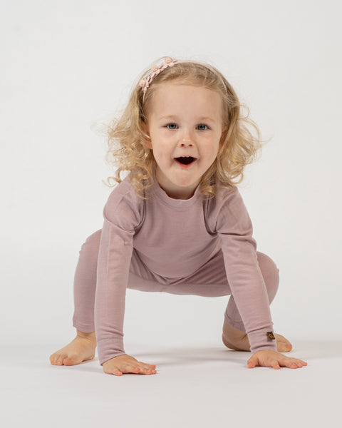 A child with curly blonde hair and a light pink floral headband is in a crouching position on a white background, wearing a dusty pink long-sleeved top and matching pants. The child has their hands on the floor and is looking at the camera with an open-mouthed expression.