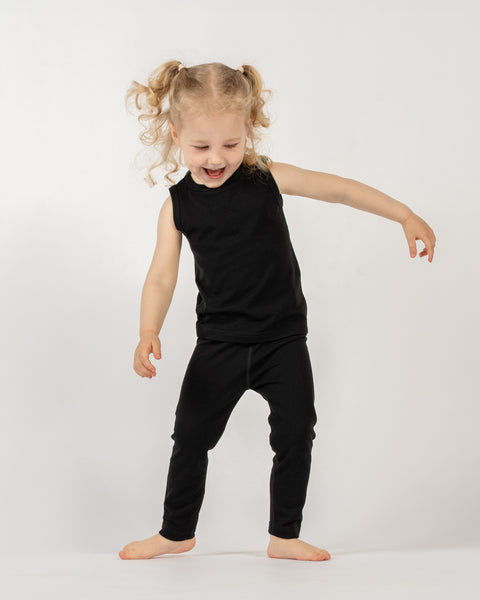 A young child, with blonde curly hair in two pigtails, is standing barefoot against a plain white background. They are wearing a black sleeveless top and black leggings. The child is captured in mid-movement, with their mouth open in a joyful expression, arms slightly outstretched, and legs bent, giving the impression of dancing or playing.