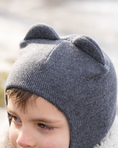 A close-up of a child's head, partially obscured, wearing a dark gray menique knit balaclava or hat with two ear-like protrusions on top. The child's right eye and part of their face are visible, and they appear to be looking slightly to their right. A textured, light-colored collar or hood of a jacket is visible around their neck, and the background is blurred with light tones, suggesting an outdoor setting.