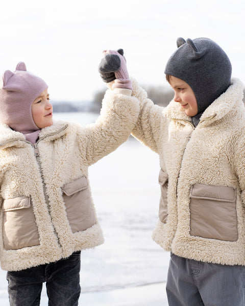 Two children, both wearing light-colored, textured jackets and menique balaclavas with ear-like protrusions, stand facing each other, raising their gloved hands to meet in a high-five. The child on the left wears a dusty pink balaclava and pink gloves, and is looking at the other child. The child on the right wears a dark gray balaclava and dark gray gloves, and is smiling at the child on the left. The background is a blurred, light-colored expanse, likely ice or water, with some distant trees.