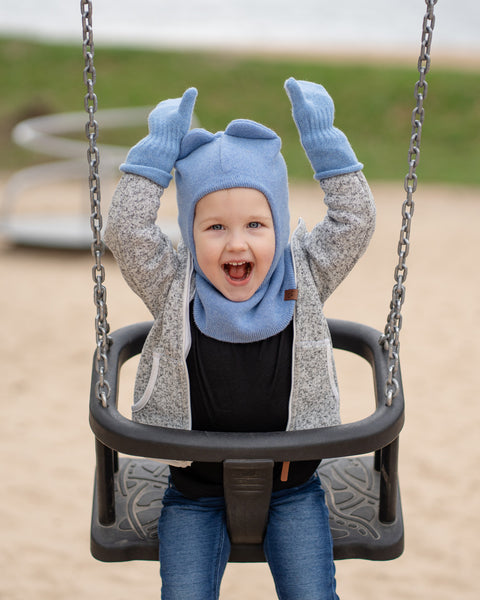 A young child is seated in a black swing seat, captured mid-motion on a playground. They are wearing a light blue knit menique balaclava with two small ear-like shapes at the top, matching light blue knit gloves, a light gray zippered jacket, a dark shirt, and blue jeans. The child has a wide-open mouth, seemingly laughing or shouting with joy, and both gloved hands are raised above their head. The background shows blurred sandy ground and some green.