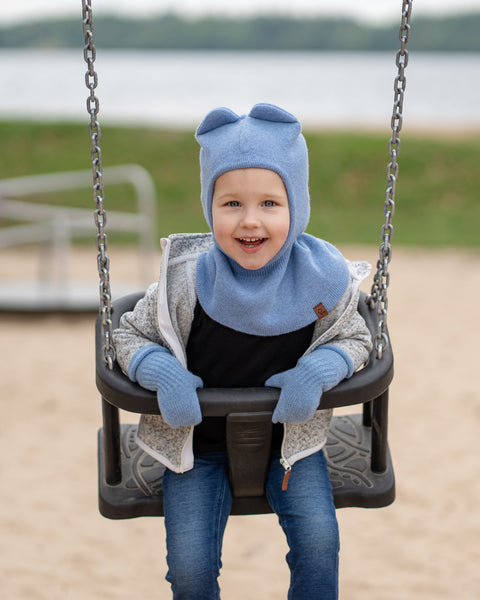 A young child, appearing to be of a young age, is seated on a swing at a playground. The child is wearing a light blue balaclava-style hat with two small, round ear-like shapes on top, matching light blue mittens, a dark long-sleeved shirt, and a gray hooded jacket with a white zipper.