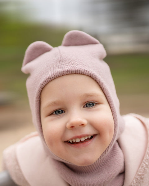 A close-up portrait of a young child with blue eyes and a wide smile, wearing a dusty pink knit balaclava with two ear-like protrusions on top. The child is also wearing a pink turtleneck or collar and a pink, textured jacket. The background is blurred, showing soft green and brown tones.