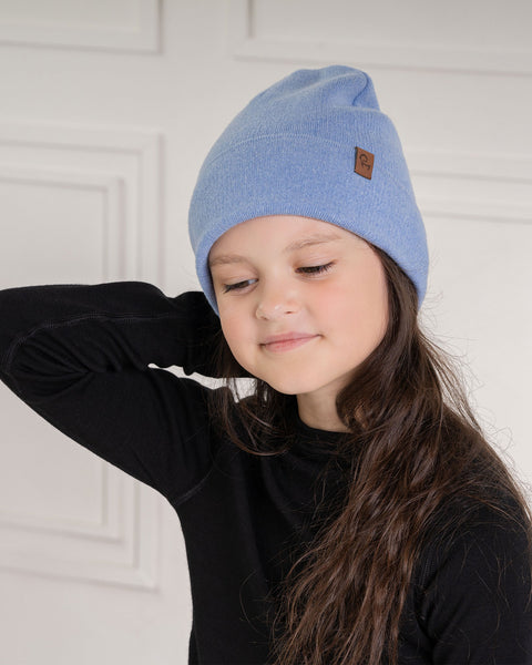 A child with long dark brown hair, is wearing a light blue knit beanie with a brown label on the side and a black long-sleeved shirt. Their head is slightly tilted down, and they are looking to the side with a soft expression. The background is a light-colored paneled wall.