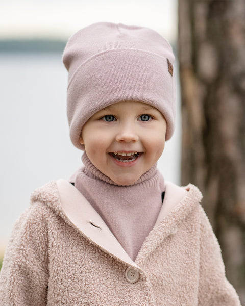 A young child with bright blue eyes and a wide, joyful smile, is looking directly at the viewer. They are wearing a light pink knit beanie, a matching pink turtleneck or neck warmer, and a beige sherpa-style coat. A tree trunk is partially visible on the right, and a blurred body of water is in the background.
