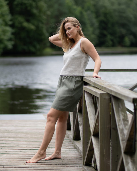 A barefoot woman stands full-length on a wooden pier, dressed in a natural linen tank top and loose stone green shorts. She leans against the railing and runs a hand through her hair, looking thoughtfully to the side against a tranquil lake backdrop.