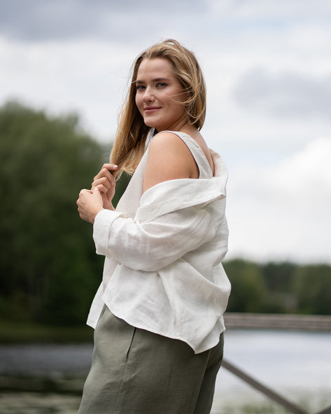 A woman stands near a scenic lake, glancing playfully over her shoulder toward the camera. Her white linen shirt is styled casually off one shoulder, paired with olive green bottoms.