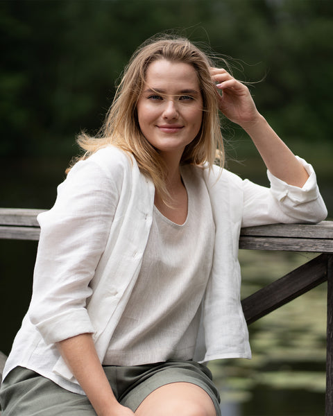 A smiling woman sits on a wooden structure wearing a natural linen top paired with stone green bermuda shorts and an open white linen shirt. She poses outdoors with a serene lake and lush green trees softly blurred in the background.