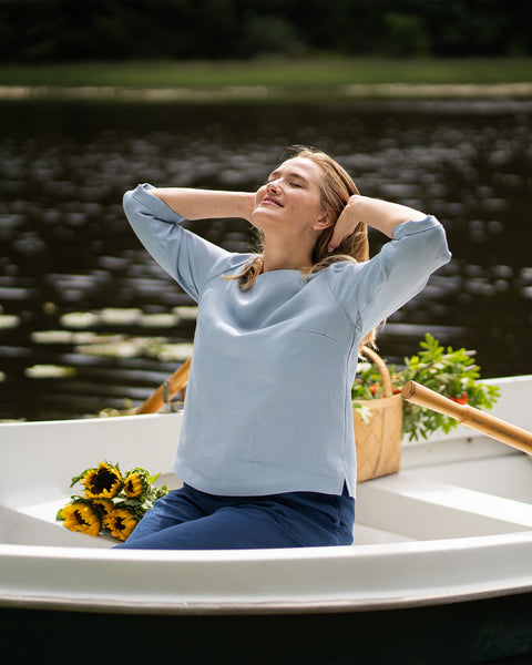 Sitting in a small boat on a calm lake, a woman stretches with her hands behind her head and her eyes closed. She wears a light blue long-sleeved top and dark blue pants, with sunflowers resting beside her.