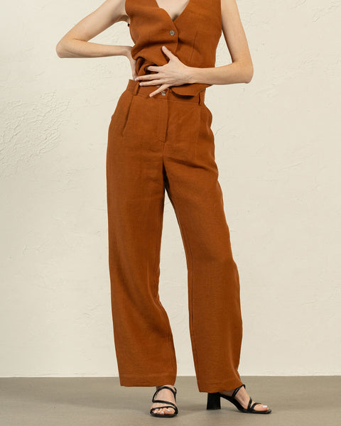 A person is shown from the mid-torso down, modeling an almond brown linen sleeveless vest and wide-legged trousers. One hand rests on their hip, while the other gently touches their stomach. They are wearing black open-toe heeled sandals. The background is a textured light beige wall and a light gray floor.