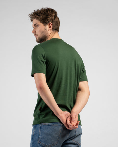 A man stands with his back partially to the camera, glancing back over his left shoulder with his hands clasped behind him. He is wearing a dark green short-sleeved t-shirt and blue jeans.