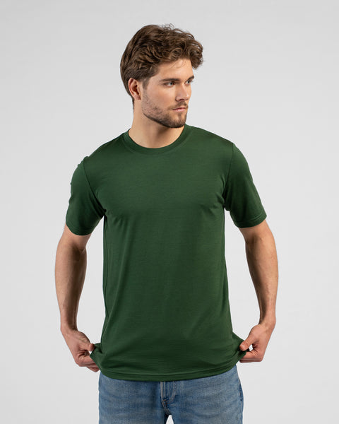 A man looks off to the right while holding the bottom hem of his dark green short-sleeved t-shirt with both hands. He is wearing blue jeans against a plain light grey background.