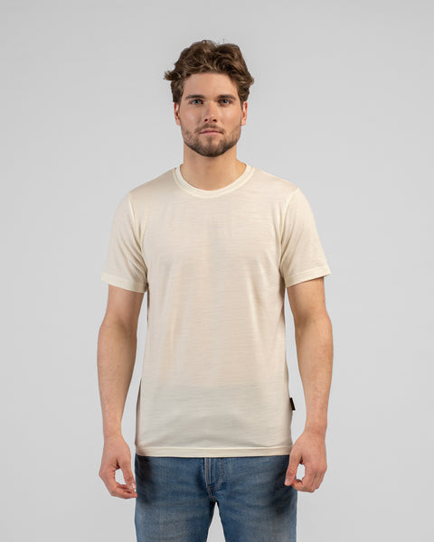 A studio portrait of a man standing and looking directly at the camera against a plain white background. He is wearing a fitted, light natural cream-colored short-sleeved crew-neck t-shirt and blue jeans.