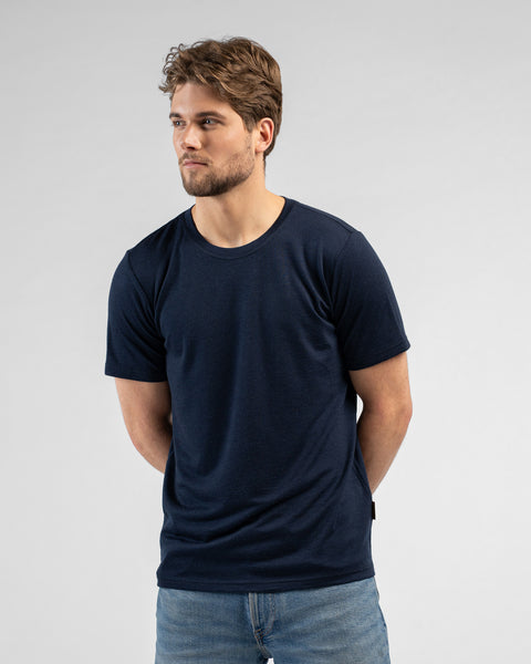 A studio portrait of a man looking off to the left side of the frame with his hands clasped behind his back. He is wearing a dark navy blue short-sleeved crew-neck t-shirt and blue jeans.