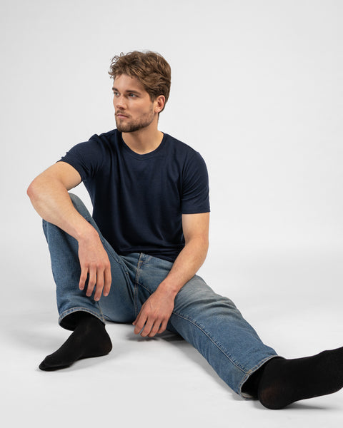 A studio shot of a man sitting on the floor with his legs extended, looking off to the left. He is wearing a dark navy blue short-sleeved t-shirt, blue jeans, and black crew socks against a white background.