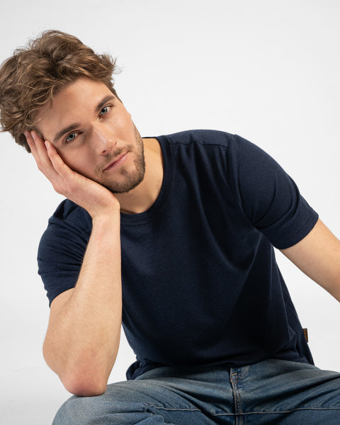A studio portrait of a man sitting on the floor and leaning forward toward the camera. He is wearing a dark navy blue short-sleeved t-shirt and resting his head on his hand.