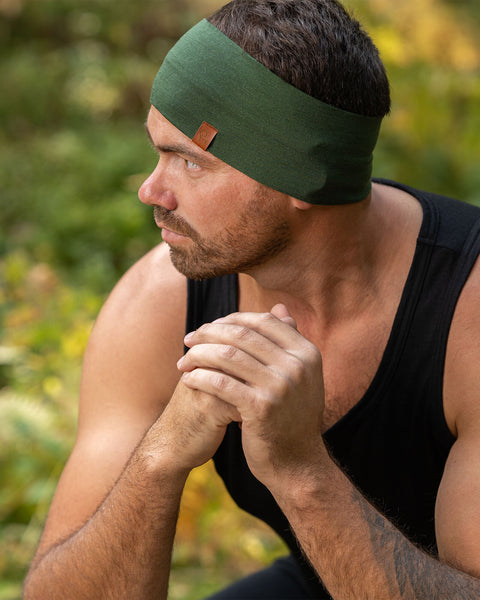 The picture features a man sitting down in a forest. He is looking to the side, with his hands on his knees, clasped together. He is wearing a dark green menique merino headbands with a small logo on the side at the front. He is also dressed in a sleeveless menique merino black top.