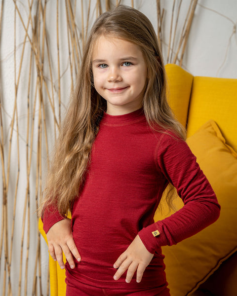 A girl wearing the menique merino long sleeve top in royal cherry. She has her hands on her hips, and is smiling at the camera. Behind her is a yellow armchair.