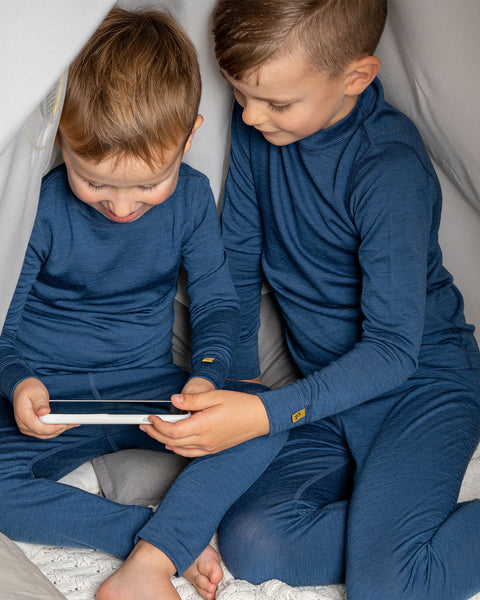 Two boys wearing the menique merino 160gsm long sleeve set in denim color. They are playing a mobile game with a phone, in a children's indoor play-tent.