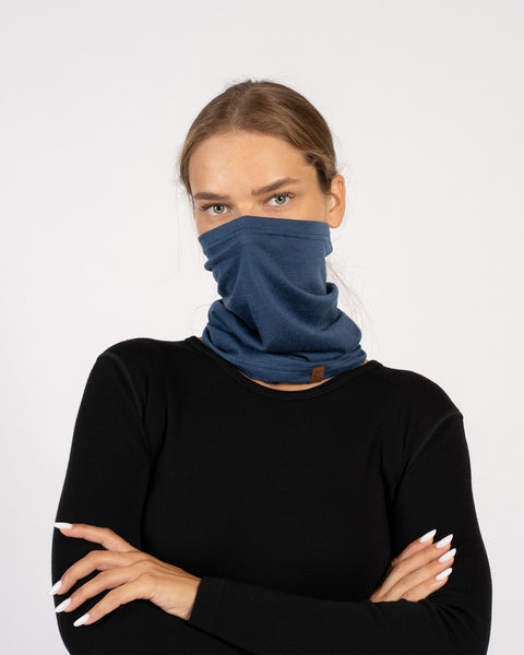 Woman with arms crossed, wearing a denim blue Merino wool neck gaiter pulled up over her nose, dressed in a black top.