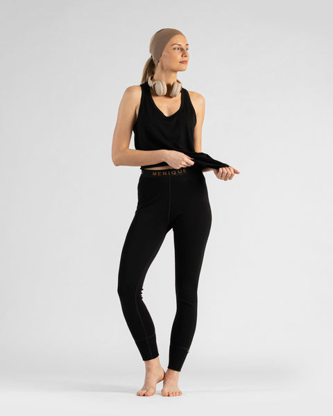 A studio portrait of a barefoot blonde woman standing and looking off to the right against a light grey background. She is wearing a black sleeveless sport top, black leggings with a "MENIQUE" logo waistband, a wide beige headband, and beige headphones resting around her neck. Her hands are lightly pulling at the hem of her top.