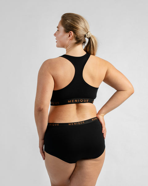 Back three-quarter view of a model looking over her shoulder with one hand on her hip, wearing a black sports bra and matching boy-short underwear, both featuring a black elastic band.