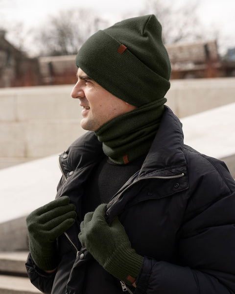 Man wearing a dark green Merino wool beanie, gloves, and neck gaiter with a black winter jacket, standing outside on a cold day.