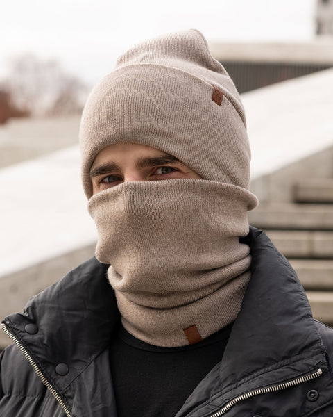 A male model, viewed in profile, wears a creamy beige beanie and a matching neck gaiter that covers his nose and mouth. Both the beanie and gaiter feature a small, brown leather patch with a Menique logo. He has a short beard and dark hair. The background is solid white.