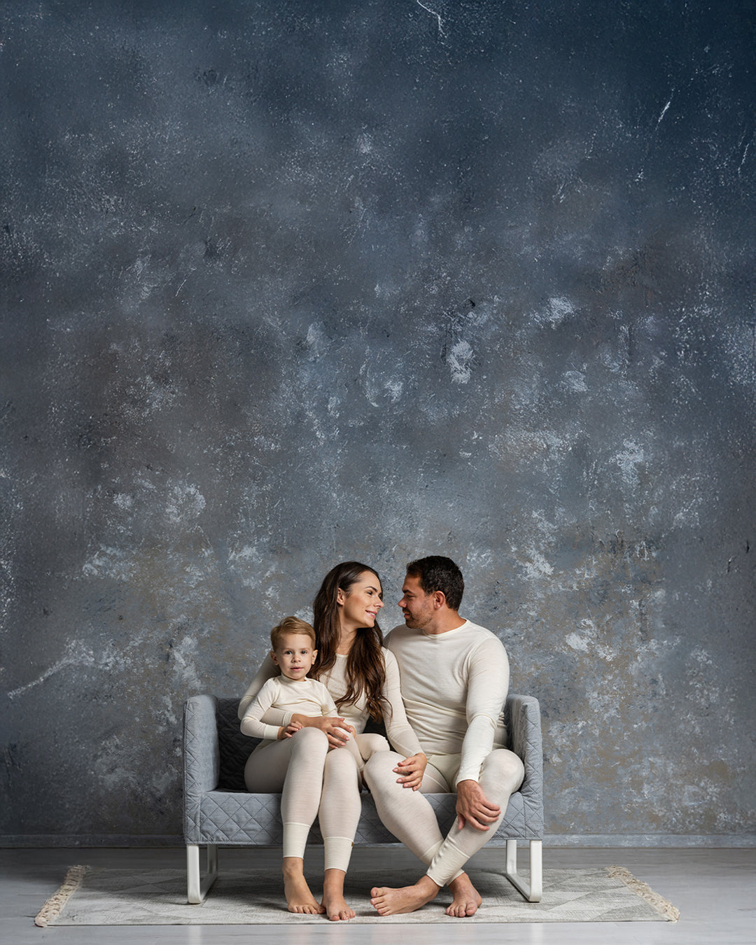 Family of three sitting on a gray sofa against a textured gray wall.
