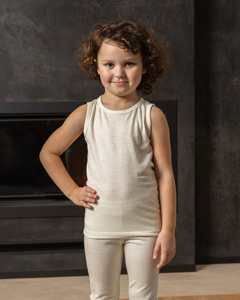 A young child with curly brown hair stands in front of a dark, textured wall with a modern fireplace opening. The child is wearing a natural sleeveless top and matching pants. They are looking directly at the camera with a slight smile, with one hand on their hip.