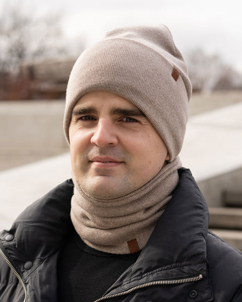 A person with light skin and dark hair is looking directly at the camera with a slight smile. They are wearing a creamy beige knit beanie with a small brown tag and a matching knit neck gaiter, also with a small brown tag. They are wearing a black puffer jacket. The background is blurred, suggesting an outdoor urban environment with concrete structures.