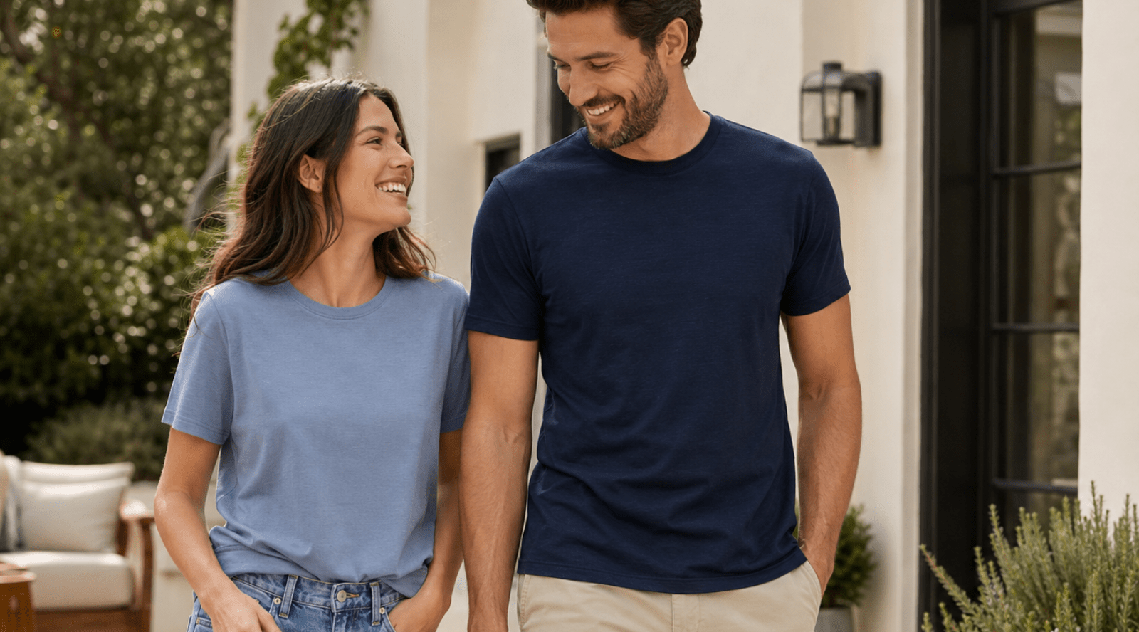 A smiling man and woman walking together outdoors near a white house. The woman is looking at the man and wearing a light blue short-sleeve t-shirt with blue jeans. 