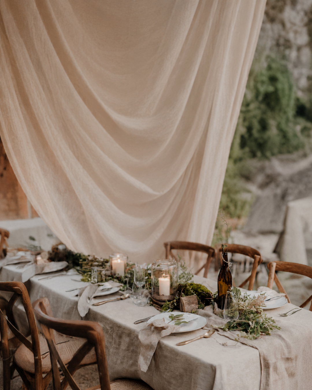 A beautifully styled outdoor dining table set for an elegant, rustic event, identical to the first image but featuring a different overhead element. Instead of foliage, a large, sweeping drape of sheer, light beige fabric hangs dramatically over the center of the table. The table is draped in a matching light linen cloth and surrounded by wooden cross-back chairs. 