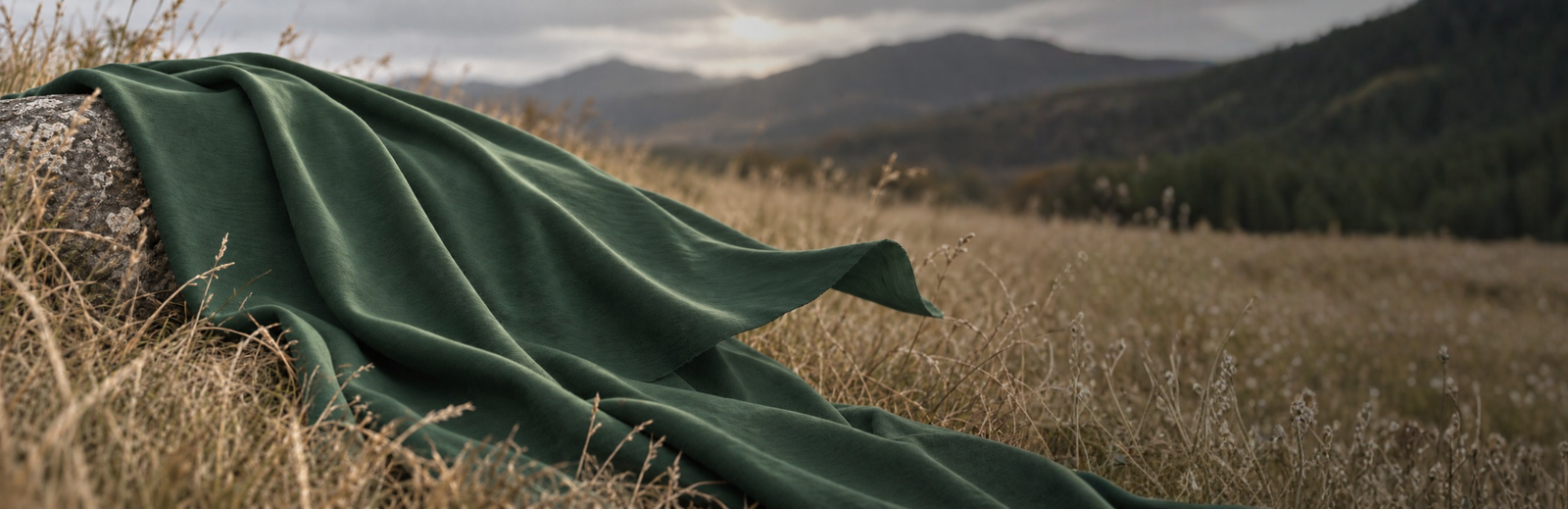 A piece of textured dark green fabric is draped over a weathered rock in a field of dry, wild grass. The fabric's edge catches a gentle breeze against a softly blurred backdrop of rolling mountains and a hazy, overcast sky.
