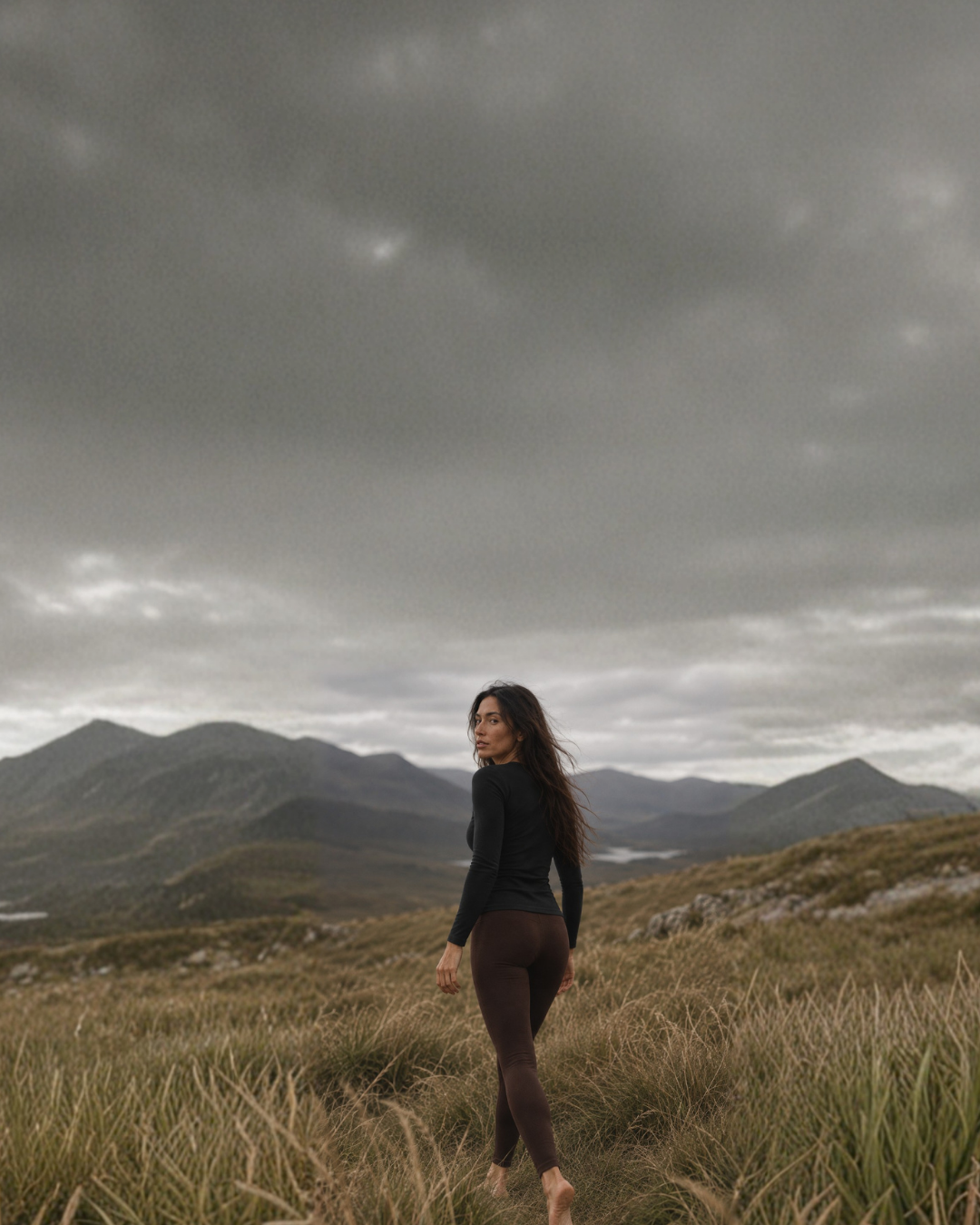 A barefoot woman with long dark hair turns to look back over her shoulder while walking through a field of tall, dry grass. She is wearing a black long-sleeved top and brown leggings, set against a mountainous landscape under a dark, cloudy sky.
