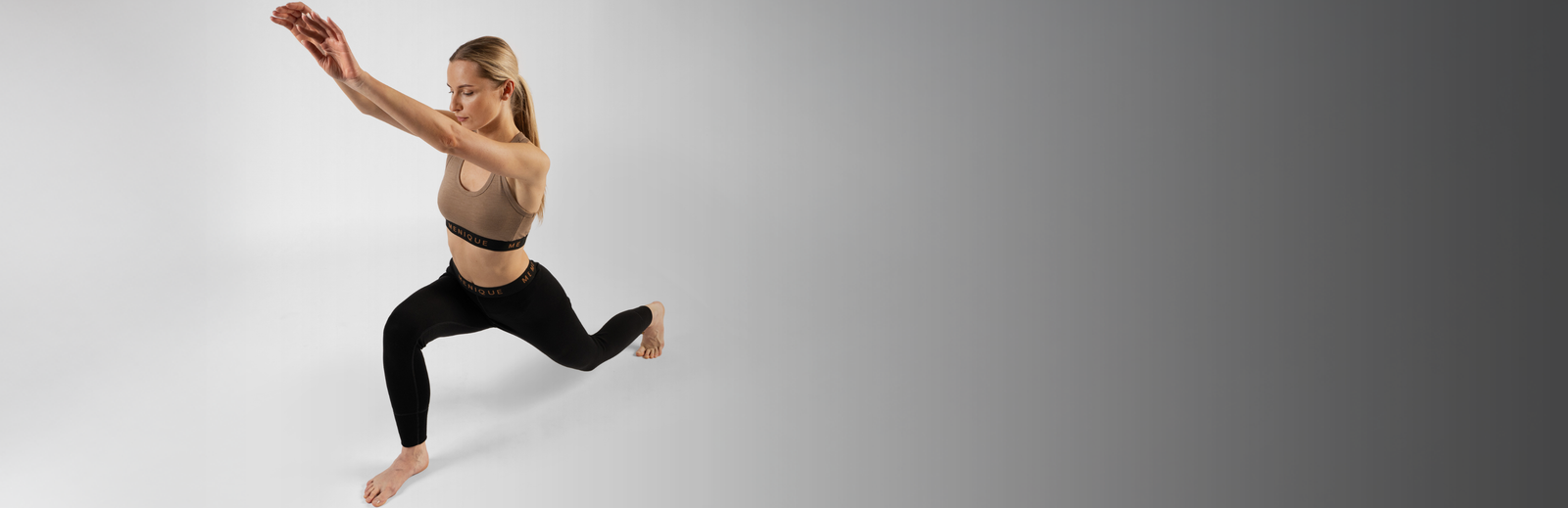 A woman in a lunging workout pose, reaching forward while wearing a brown sports bra and black leggings, both featuring Menique branded waistbands, set against a solid grey background.