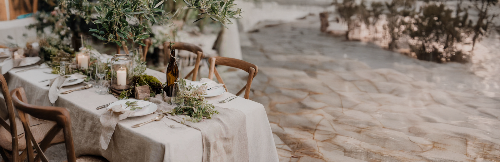 A beautifully styled outdoor dining table set for an elegant, rustic event. A dramatic, lush branch of green foliage, resembling olive branches, overhangs the center of the table. The table is draped in a light, natural-colored linen cloth and surrounded by wooden cross-back chairs. The place settings include white plates, folded linen napkins, and gold flatware. 