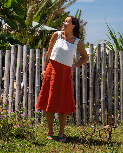 A woman with dark hair pulled back in a ponytail, wearing a pure white sleeveless crop top and menique midi skirt, stands with one hand on her hip and looks upwards and to the left, smiling, in front of a light-colored bamboo fence and lush green tropical foliage. She is wearing patterned sandals. The setting appears to be outdoors on a sunny day.
