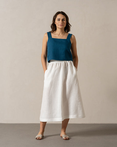 A woman with medium-toned skin and dark, shoulder-length hair stands facing forward against a plain, light-colored wall. She is wearing a teal, sleeveless top with a square neckline and a white, menique skirt with her hands in the pockets. Her feet are bare, and she appears to be standing on a light gray floor.