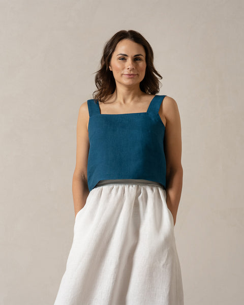 A woman with medium-toned skin and dark hair looks directly at the camera with a neutral expression. She is wearing a teal, sleeveless square-neck linen top and a pure white, menique
 linen skirt, with her hands in the skirt&#39;s pockets. The background is a plain, light beige color.