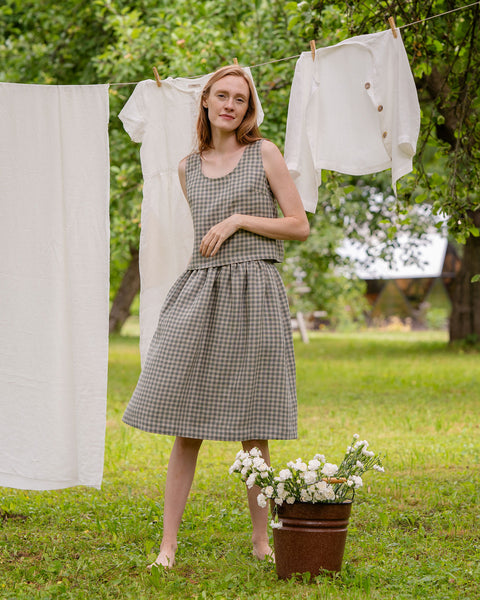 In this picture the model is standing in front of a clothesline with white garments hanging on it. Beside her is a metal bucket full of flowers. She is visible full-height, as well as the full skirt length. The skirt is knee length, matched with a same pattern blouse.