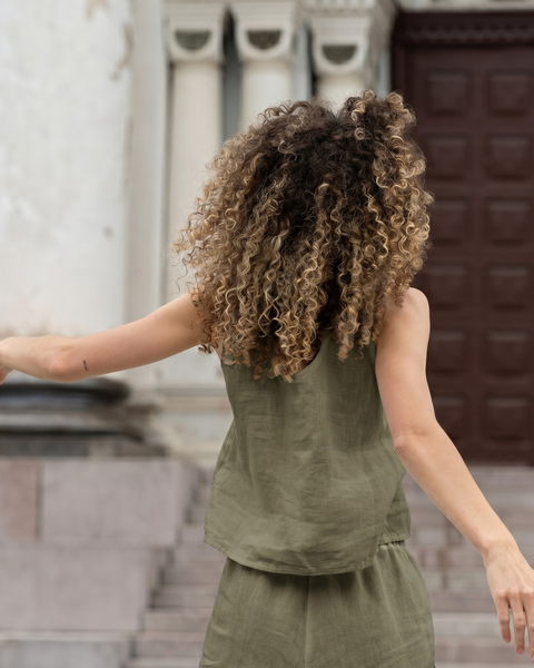 A woman with voluminous curly hair viewed from behind, wearing an olive green sleeveless linen top and matching pants. Her arms are slightly outstretched, and she is standing in front of wide stone steps leading up to dark wooden doors.