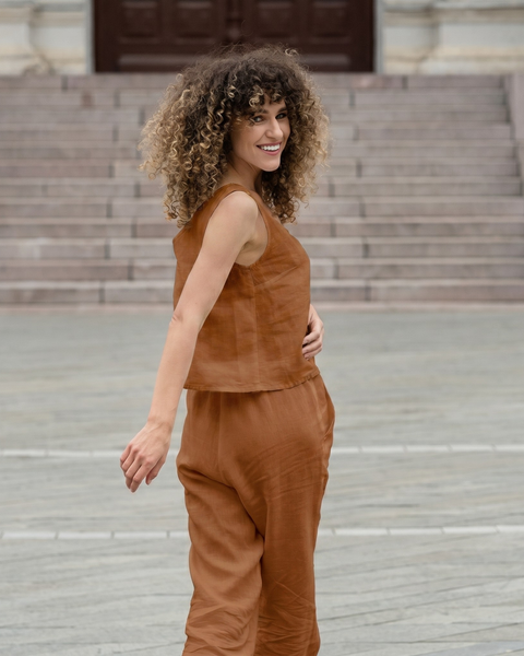 A woman with curly hair looking over her shoulder with a smile as she walks away across a stone plaza. She is wearing a rust-colored, almond brown two-piece linen outfit.