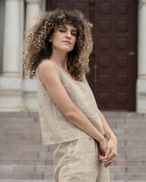 A woman with curly hair standing in front of stone steps, looking confidently at the camera. She is wearing a light beige, natural-colored linen sleeveless top and matching pants, with her hands resting gently near her waist.
